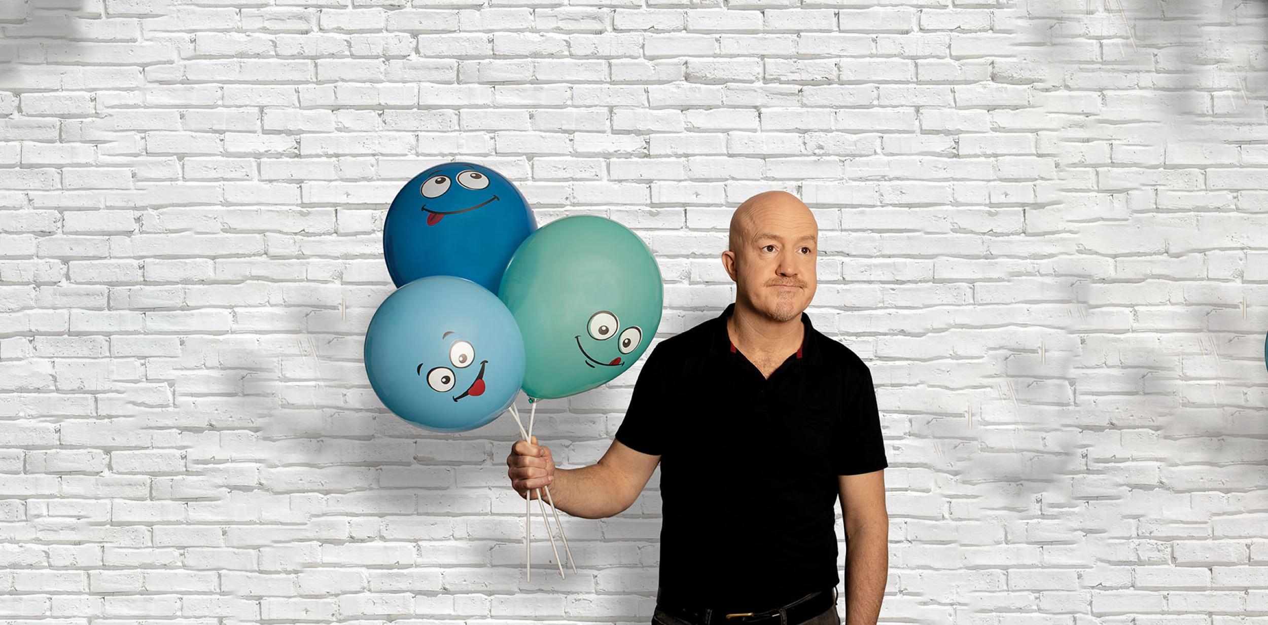 Andy Parsons in black tshirt holding 3  blue balloons with  illustrated faces on them,  against a white wall backdrop