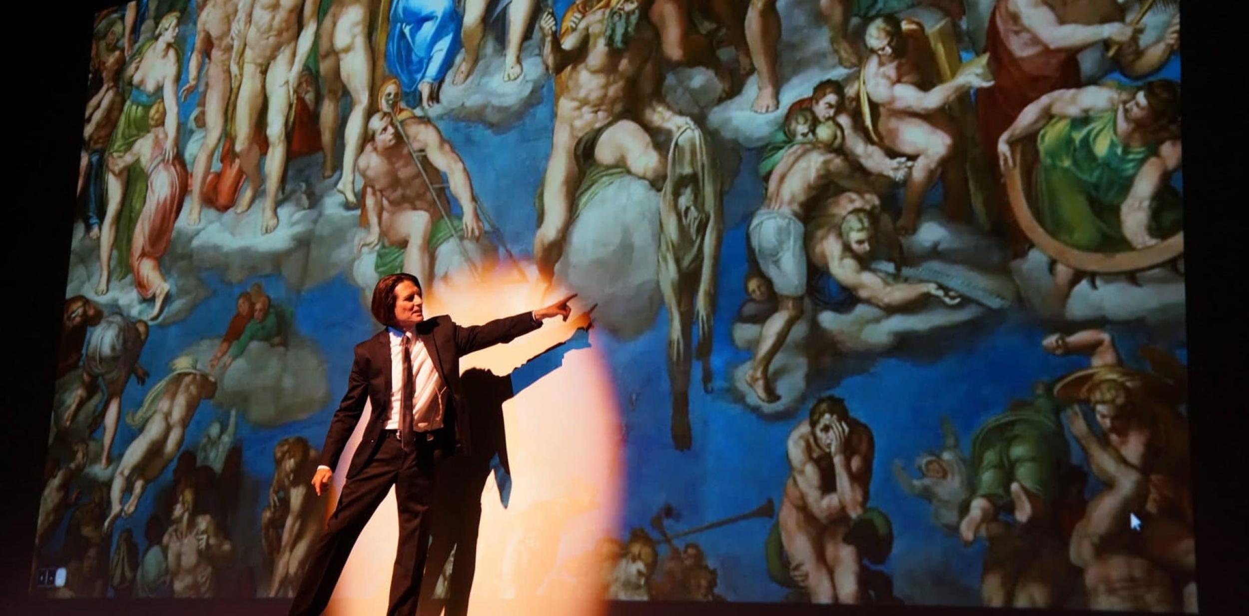 A person on stage pointing at a large piece of classical artwork