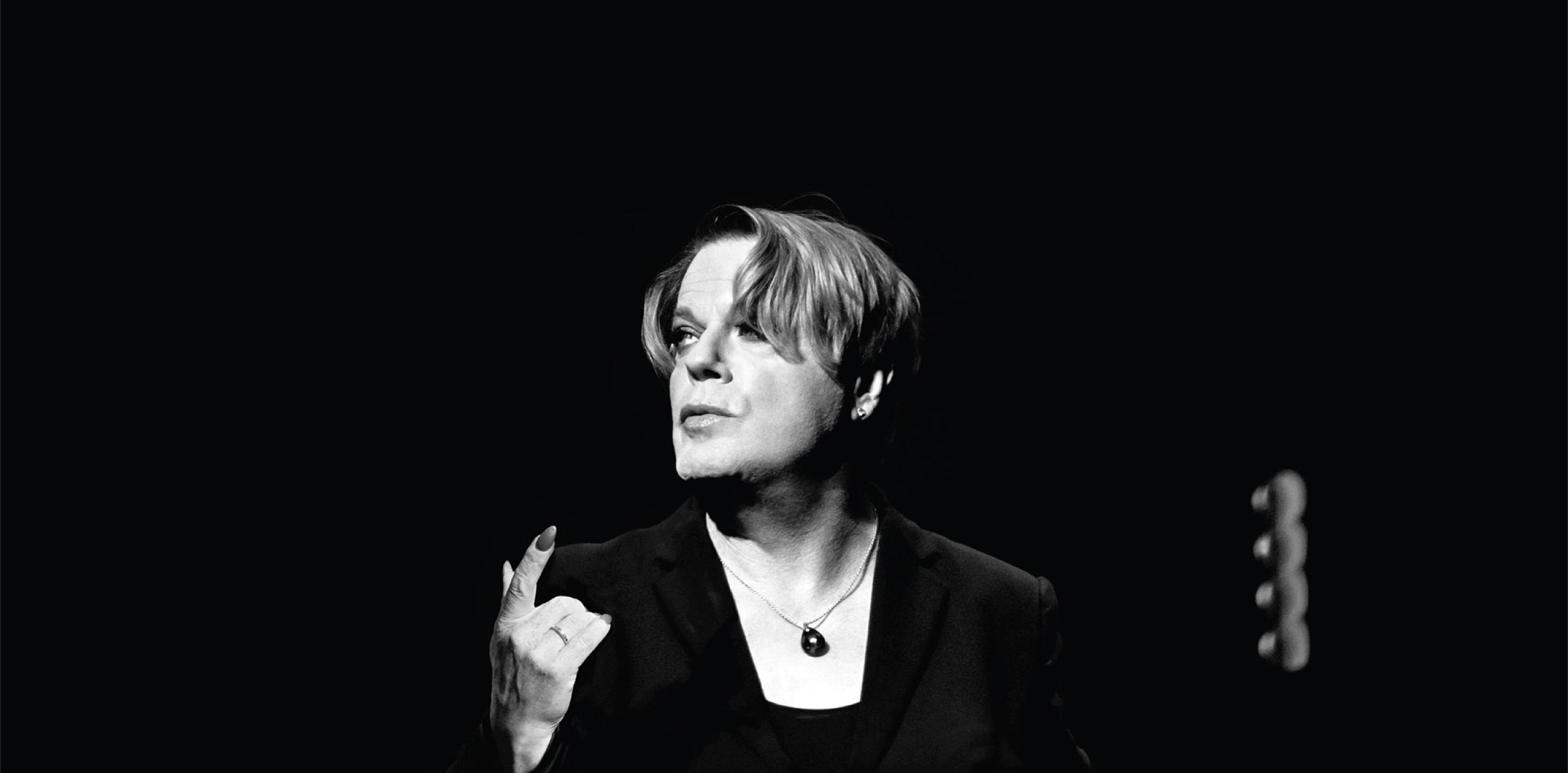 Black & white image of Eddie Izzard looking up to the left pointing their finger up