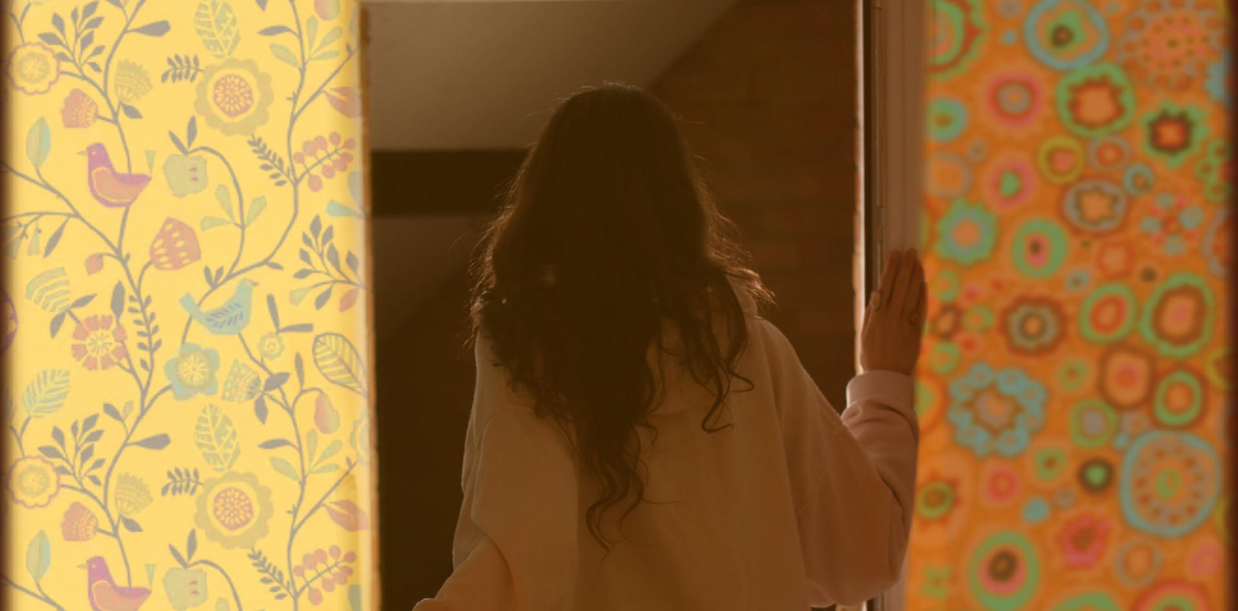 Young girl with back to us, looking through a door with 70s wallpaper either side