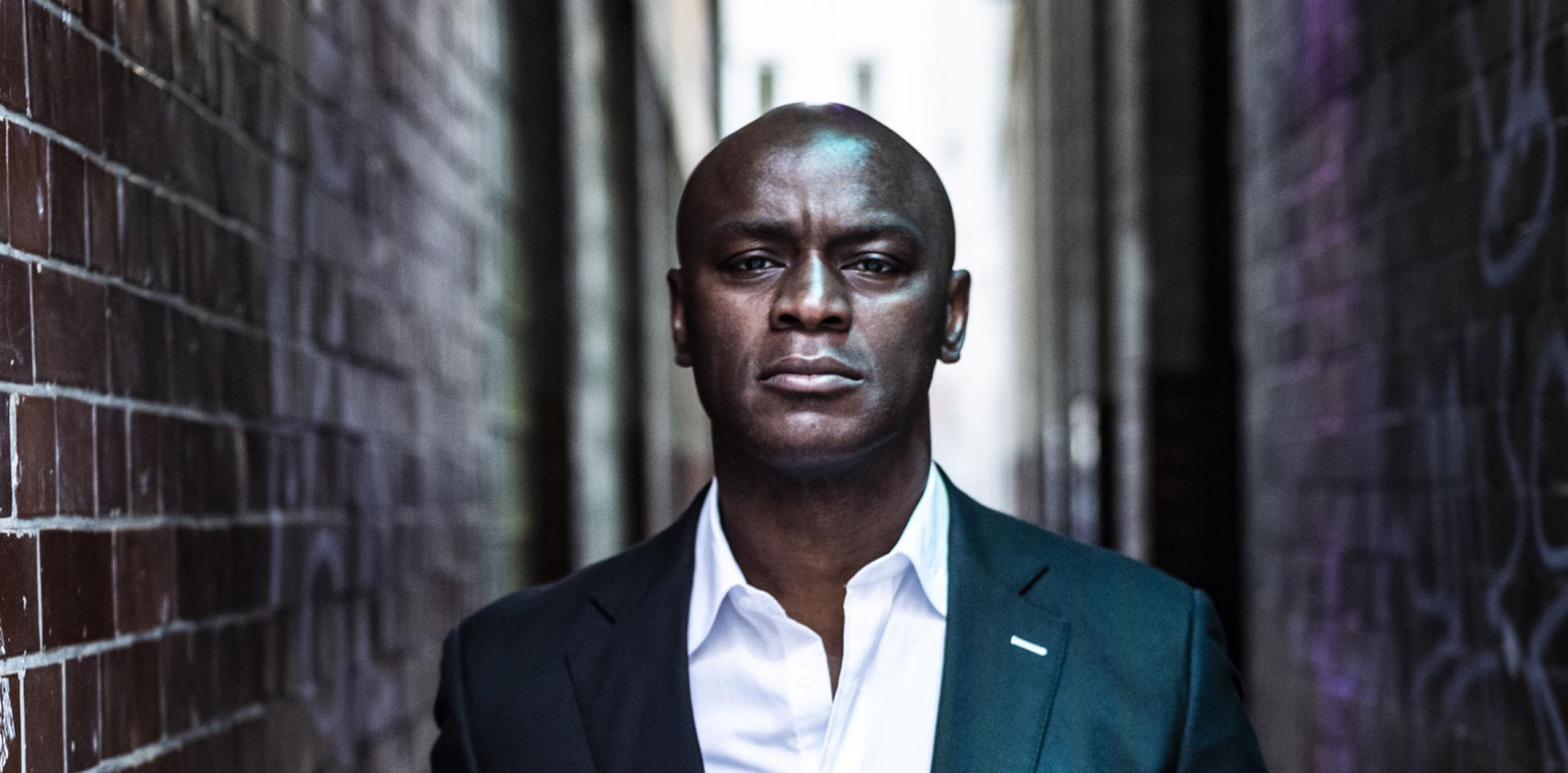 Ola Onabule in a suit , but tieless, walking down an alleyway looking at the camera