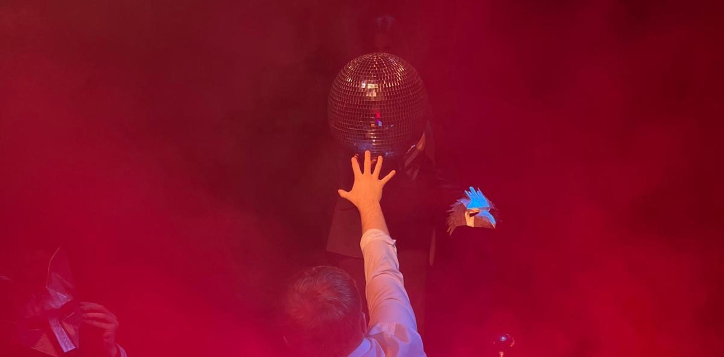 Red hazy image of a man with his back to the camera, reaching out to touch a mirror ball