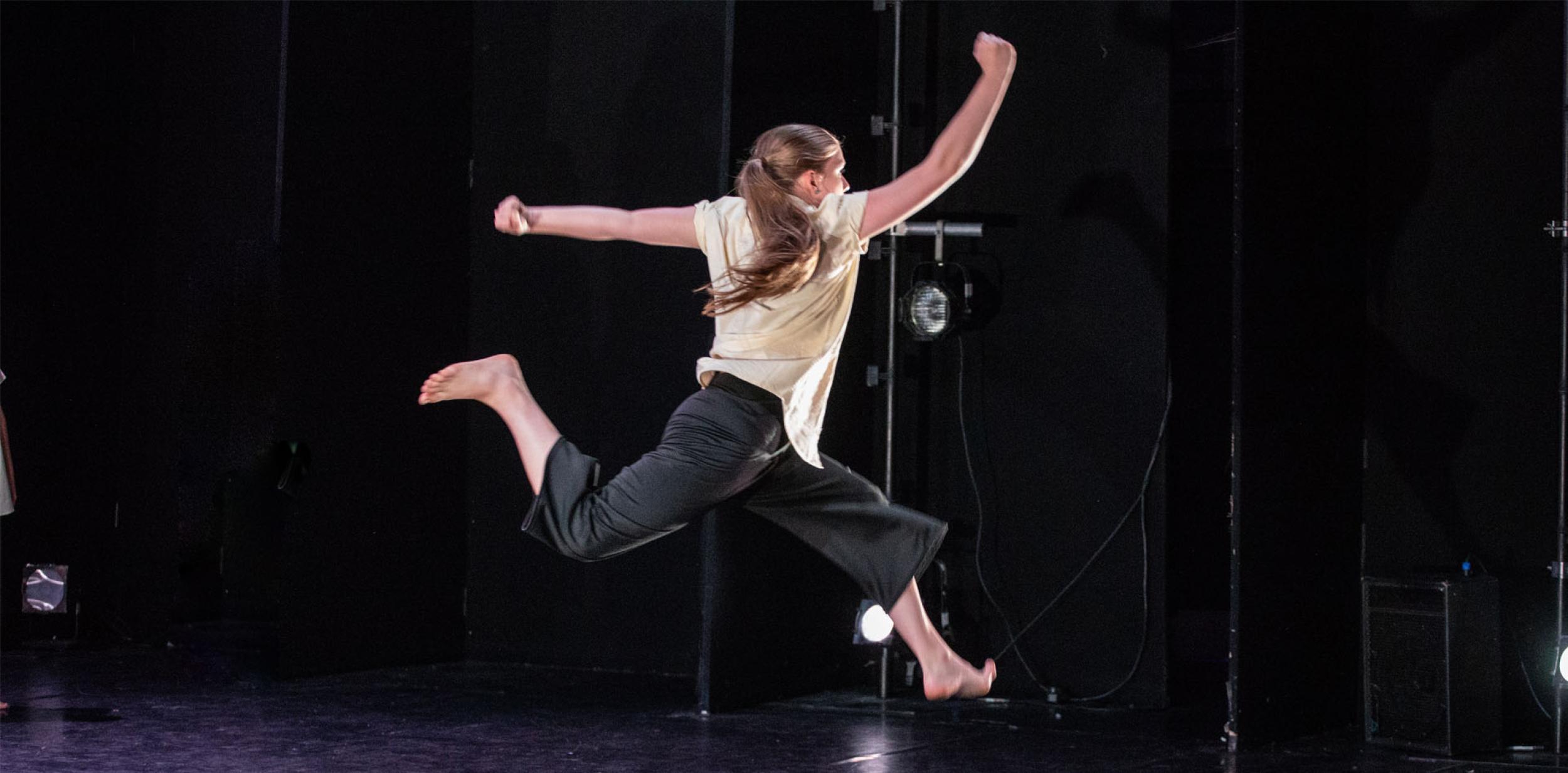 Image of young female dancer in black loose trousers and white T-shirt leaping on a black stage