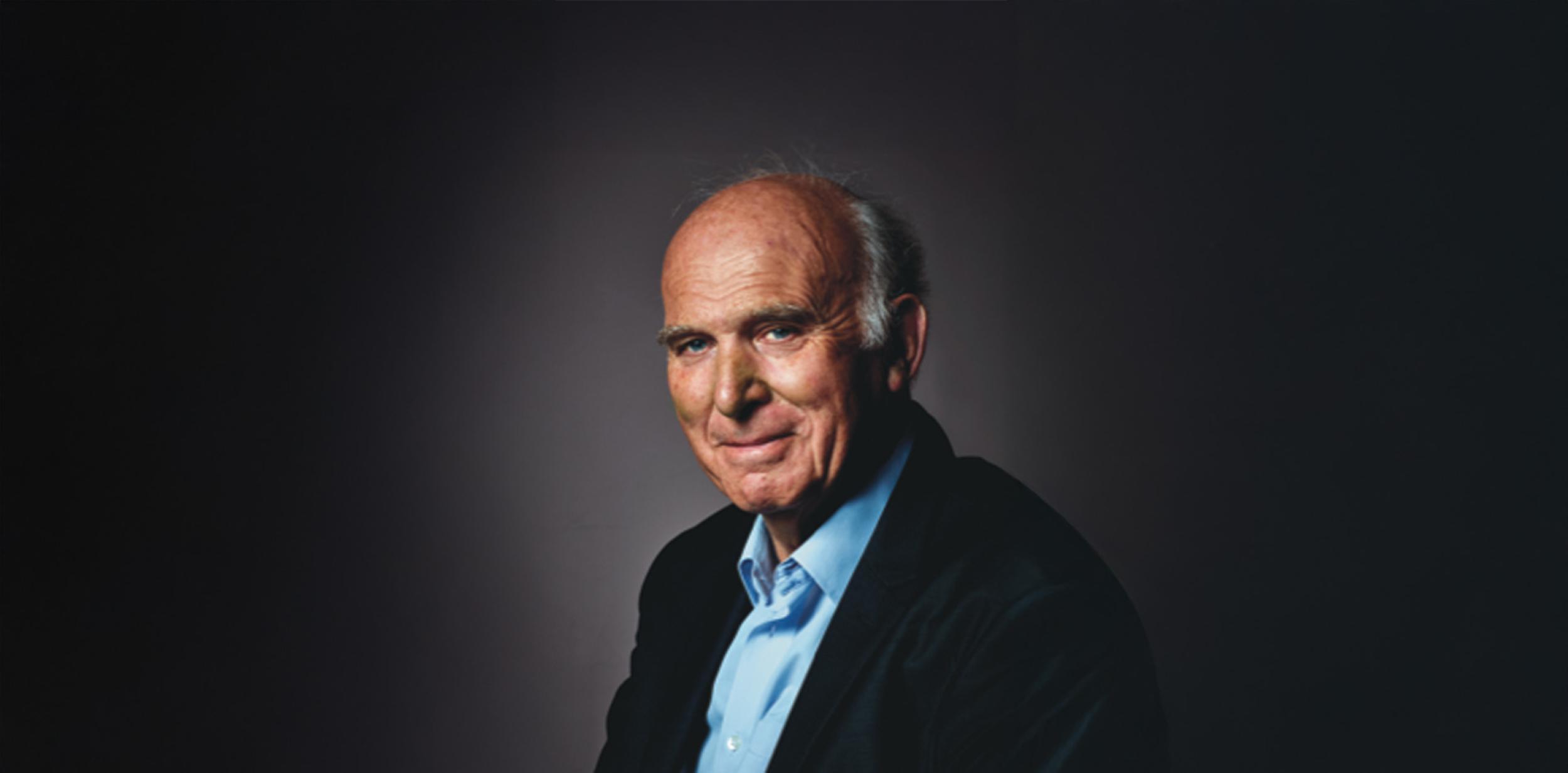 Vince Cable in blue shirt, blue suit jacket in front of a plan dark background, looking at camera