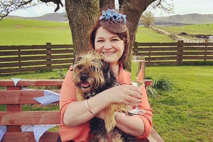 Gill Sims sat outside in countryside holding a dog and a glass of champagne