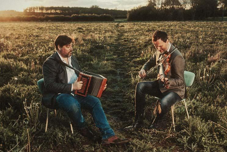 Two men playing musical instruments sat on chairs in a field