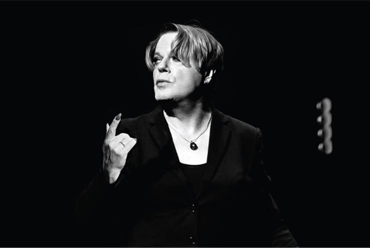 Black & white image of Eddie Izzard looking up to the left pointing their finger up