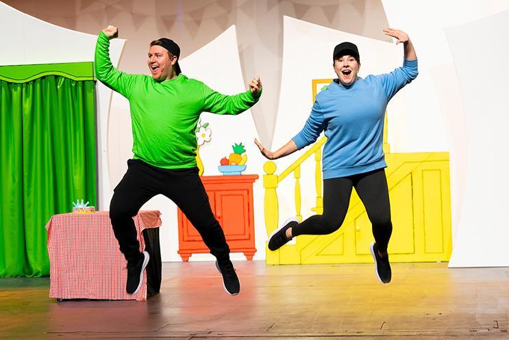 2 actors leaping in the air, 1 wearing a bright green jumper, the other a bright blue one