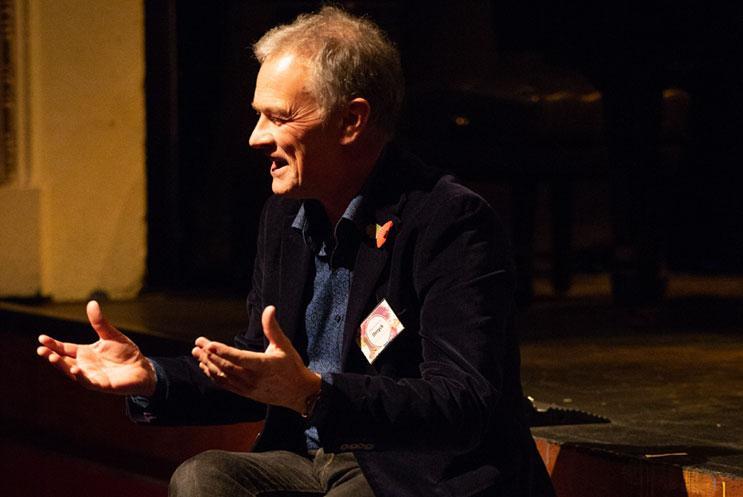 A man sat on a stage giving a speech and gesturing with his hands